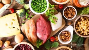 Variedad de alimentos ricos en proteínas sobre mesa rústica, incluyendo carne, pescado, queso, legumbres y frutos secos