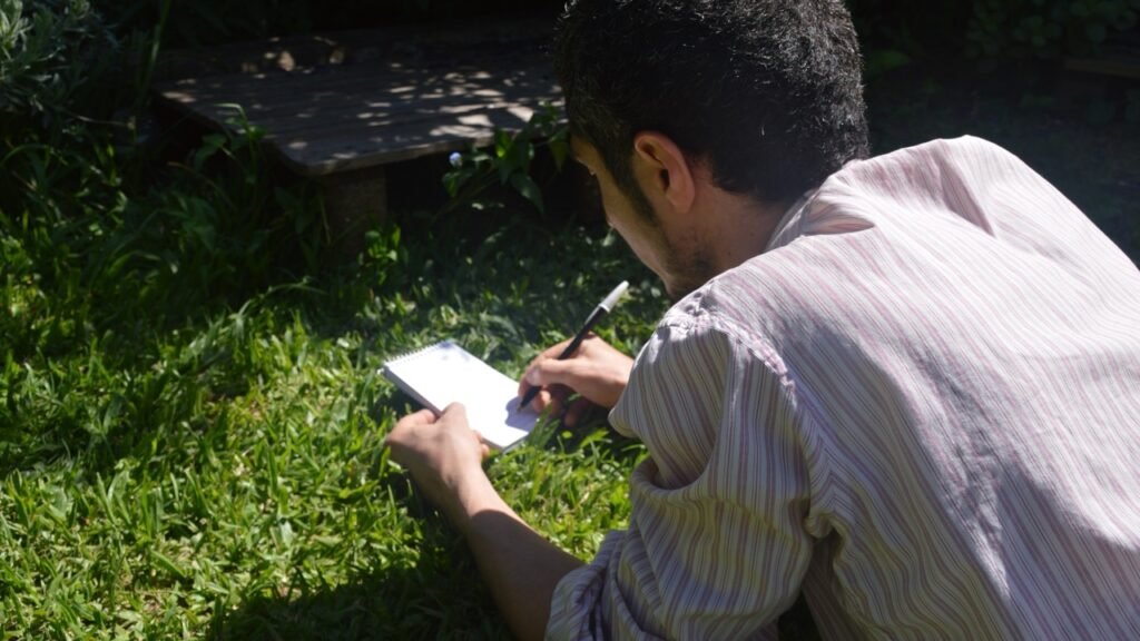 Hombre escribiendo en un cuaderno mientras se encuentra acostado en el pasto al aire libre. Fotografías de Ximena Frabegat